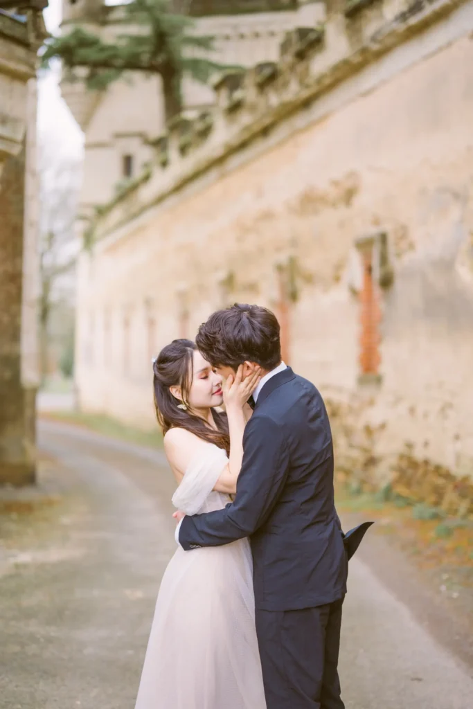 Noorpaar pulmarõivas embab lossihoovi tänaval. Peigmehe käsi toetub neiu puusale ja pruudi käed on peigmehe põskedel. Nad on suudlemas. 