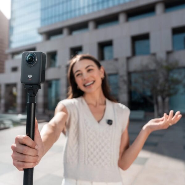 DJI Osmo 360 kaamera on selfipulga otsas, mida hoiab tumedapäine noor neiu, kes kannab valget lühikeste varrukatega pluus. Ta seisab linnatänaval ja õues on päikeseline ilm. Tema näol on naeratus.