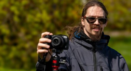 Imelise rohelise looduse taustal seisab päikeseprille kandev meesterahvas. Ta kannab habet ja tal on pikemad juuksed. Tema parem käsi on sirutatud välja Nikon Z30 kaamera päästikule. Ta teeb timelapse. Kaamera on asetatud statiivile.